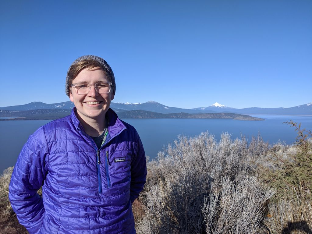 person in a blue puffy coat and stocking hat, standing atop a hilltop with a giant lake in the background