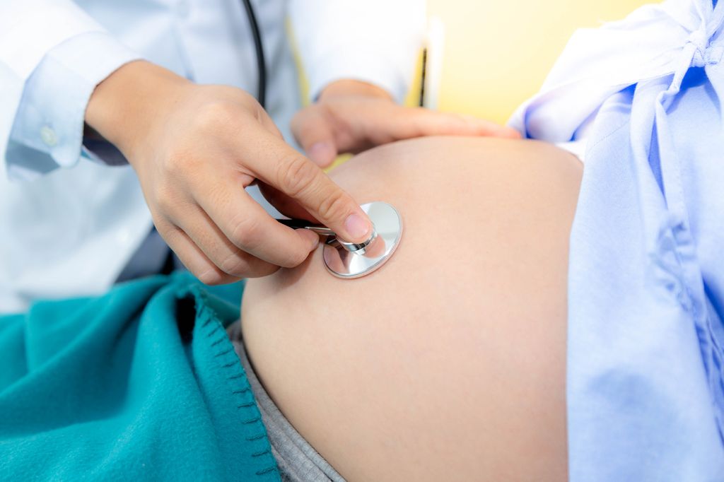 Beautiful pregnant woman is lying down on a bed at hospital room. Handsome doctor is checking fetus heartbeat and fetus position at mother belly by using stethoscope. She nearly gives birth next month
