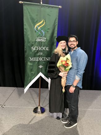 woman in green cap and gown, with her husband, in front of a sign for OHSU School of medicine graduation