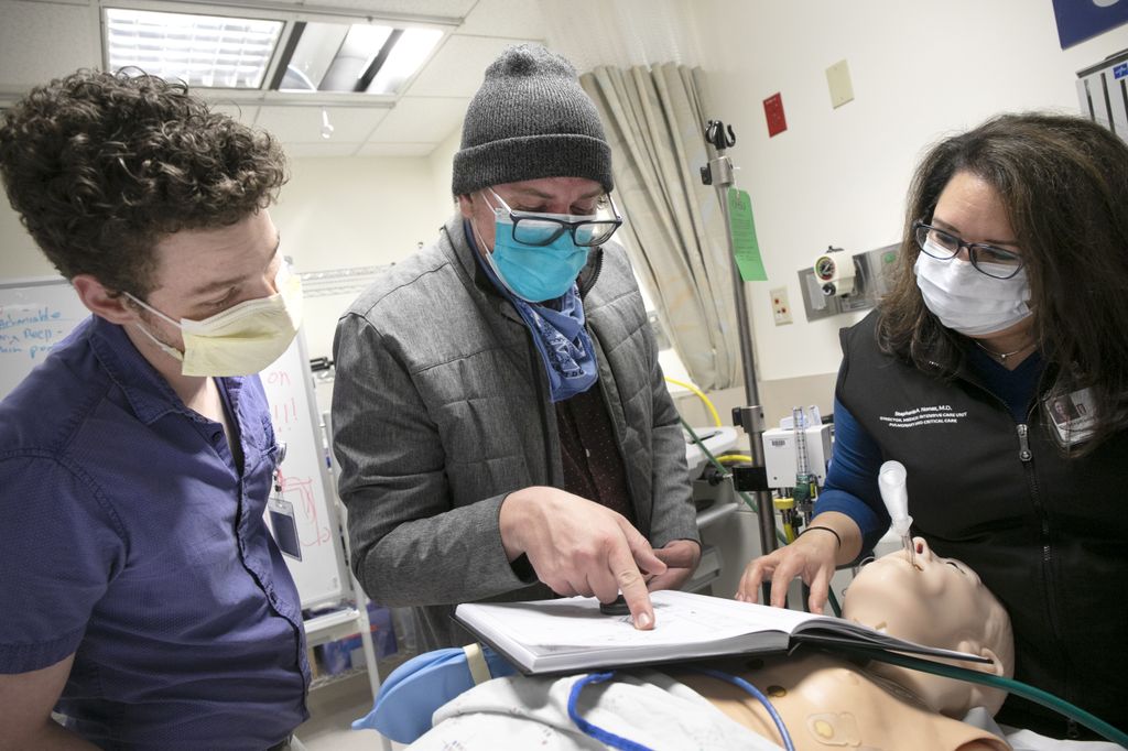 three people standing bedside near a training dummy, looking at book together.