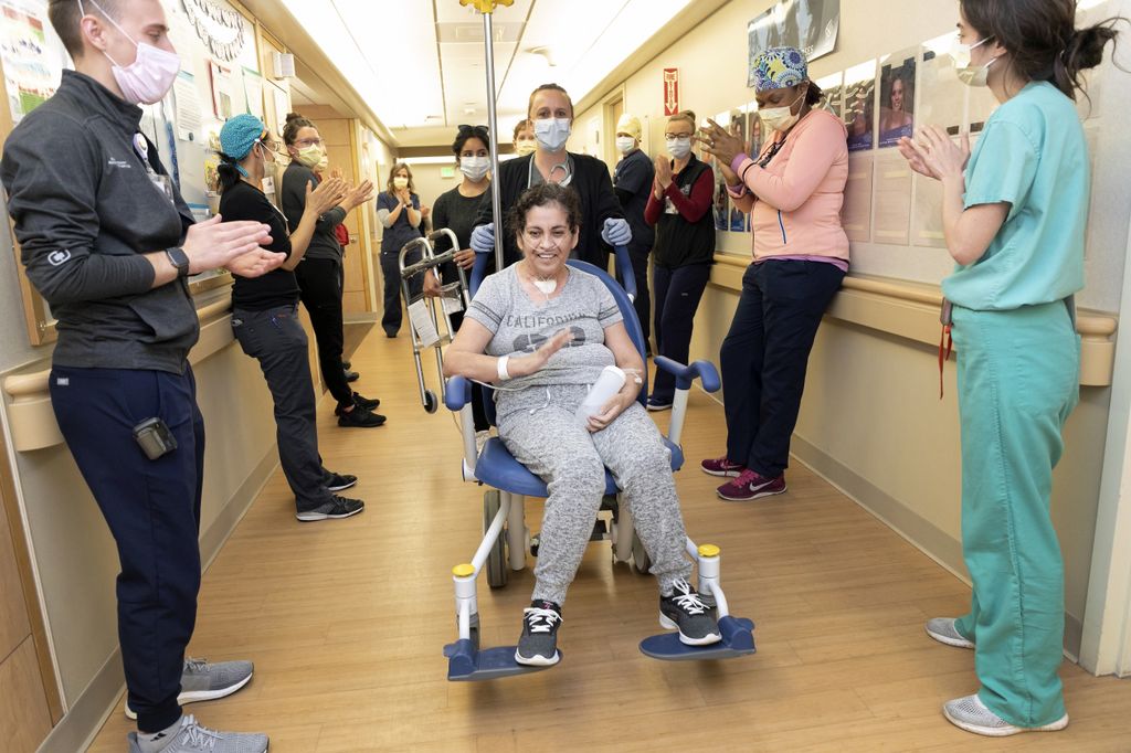 woman being wheeled out of a hospital down a hallway filled with people