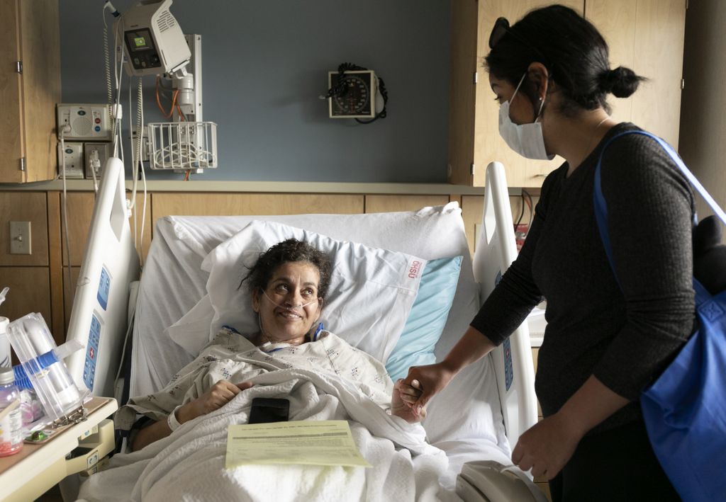 woman in her hospital bed holding her daughters hand and looking up to her