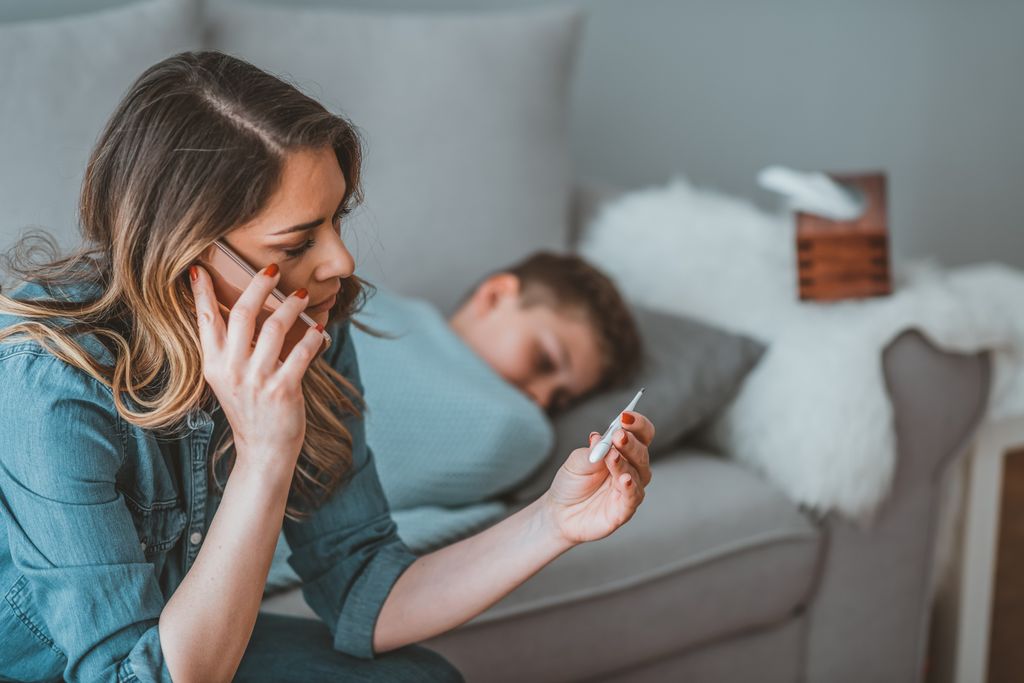 mother sitting on couch, on the phone, reading the temperature of a child who is covered with a blanket