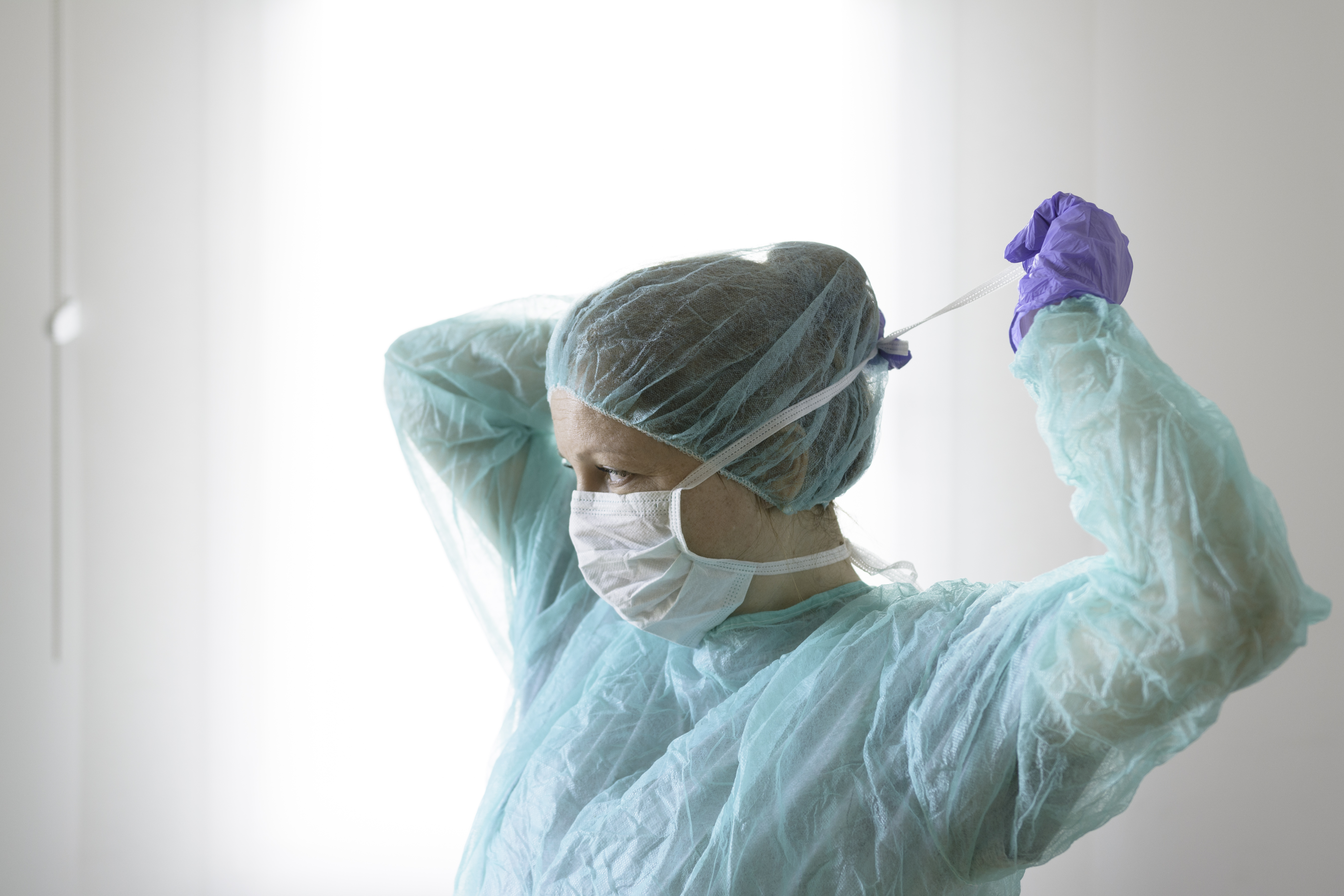 a woman with her hair covered in a surgery scrub cap, wearing greening surgery material, tying on a face mask