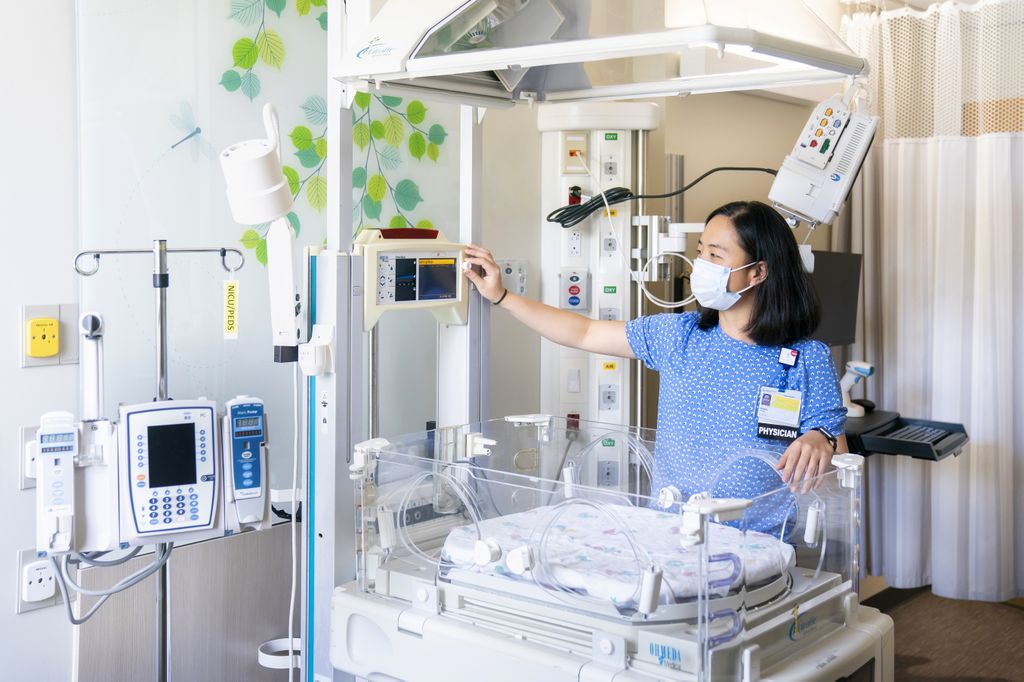 doctor in a light and bright room, which is a NICU, adjusting a monitor over an empty infant bed