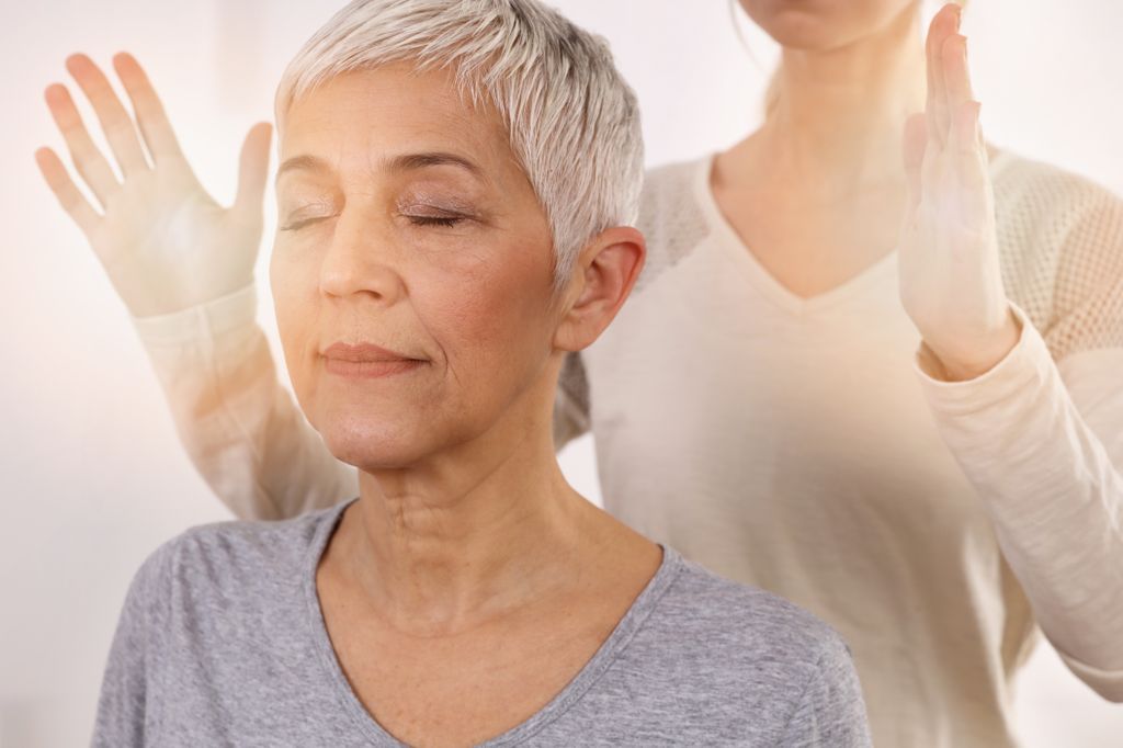 a woman, shown from shoulders up, with a second woman standing behind her, hands raised, as she begins a reiki treatment