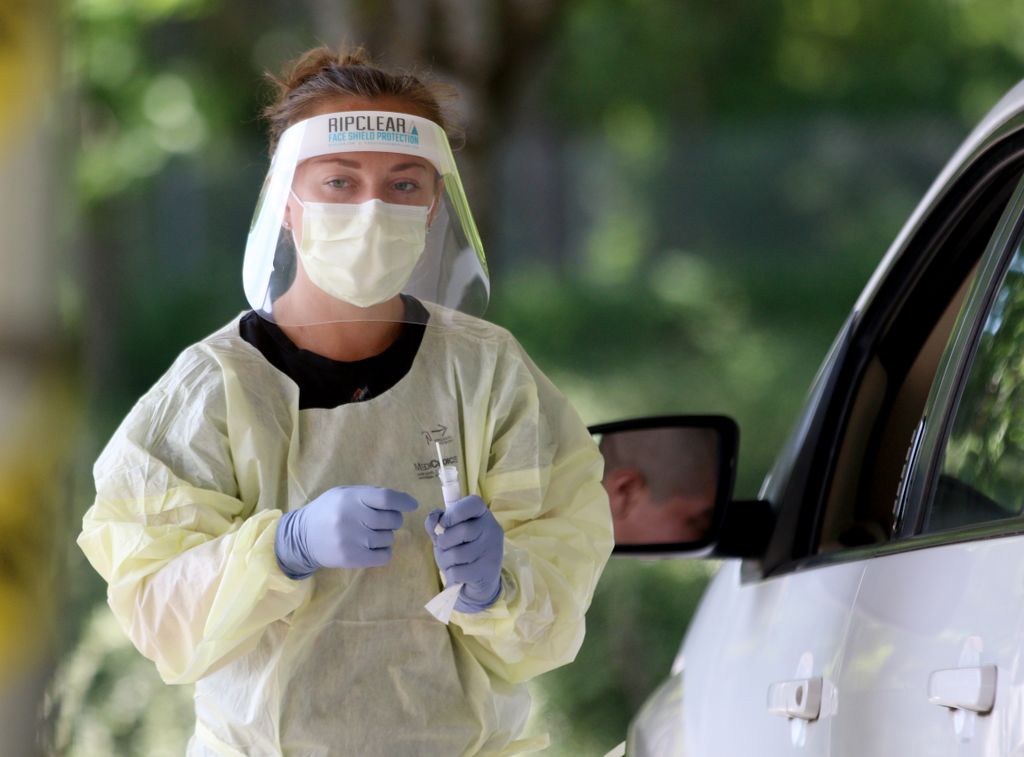 A woman wearing surgical gloves, a protective gown, face mask and face shield stands near an open car window