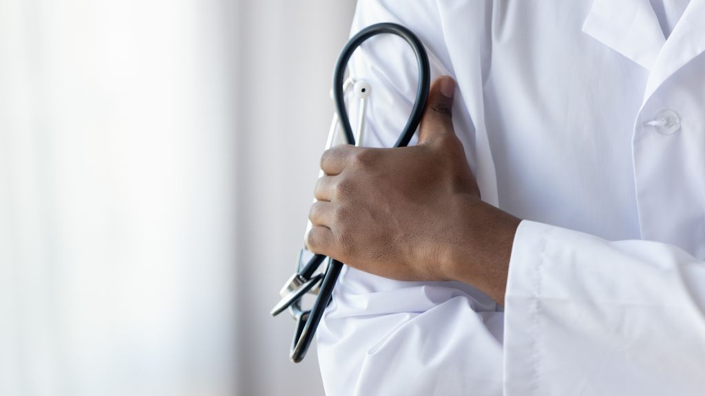 close up shot of a black man's hand, holding a stethoscope, wearing a white coat
