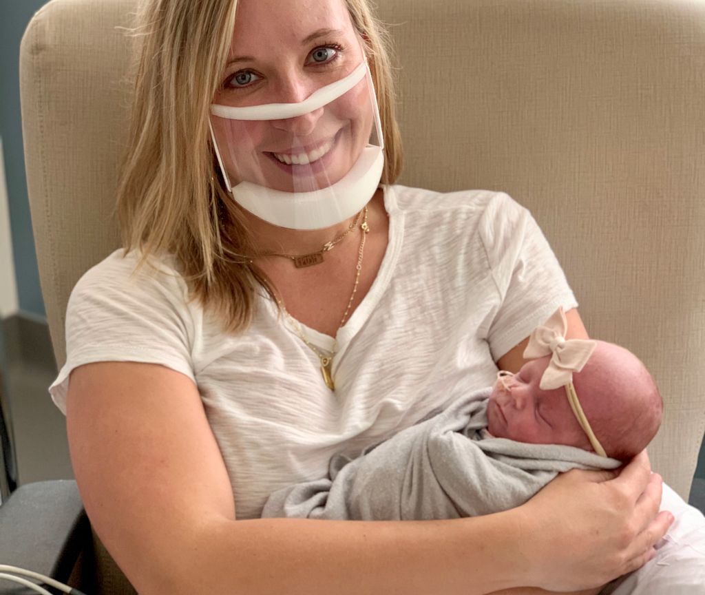 A mother holds her infant daughter, with her smile visible through a clear mask.