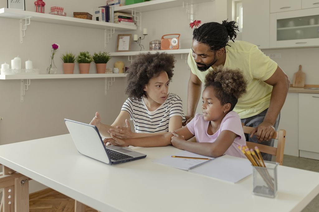 Black parents with their daughter, working on distance learning at a table with a laptop, pencils and notebook