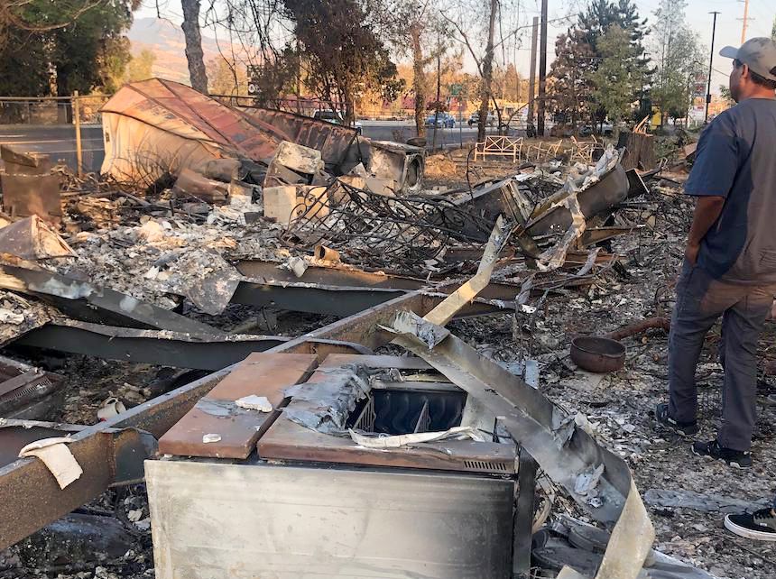 burnt out remains of a home following a wildfire, man standing to the right, seen from back, looking over remnants of the home