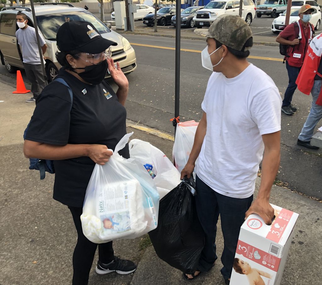 A masked female volunteer gives a box of diapers and bags of clothing to a masked man.