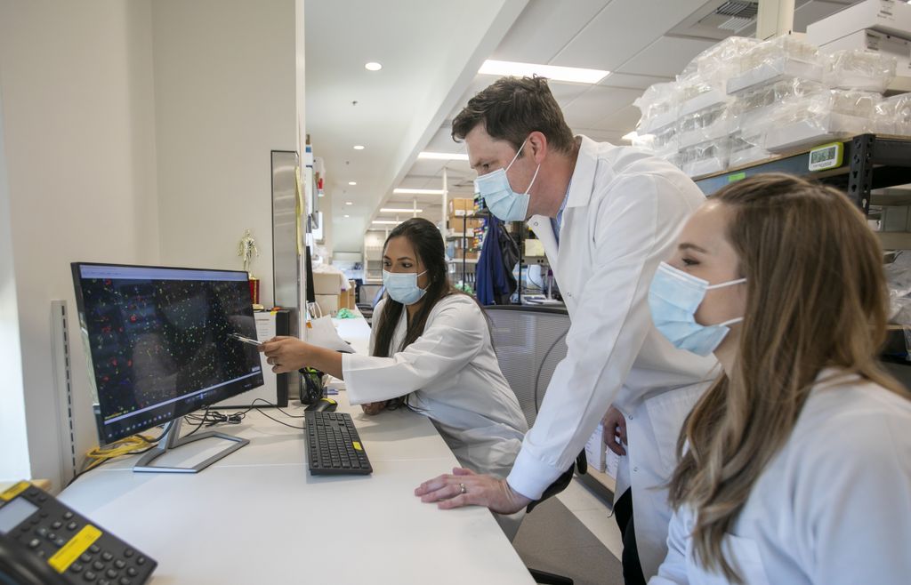 Ben Burwitz, a white male in a lab coat, with two female colleagues, Lauren Rust and Sreya Biswas, looking at a computer monitor on a desk in a medical lab.