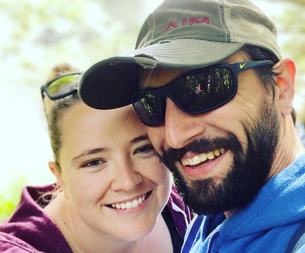 A close-in shot of a couple: Catherine Adams (left), a smiling woman with her hair pulled back; and her fiance (right), a smiling, bearded man in sunglasses and a ball cap