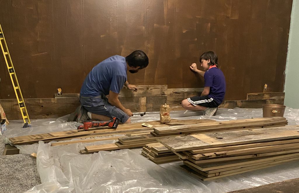 A man and boy work together on finishing a dark brown accent wall; stacked planks are in the foreground.