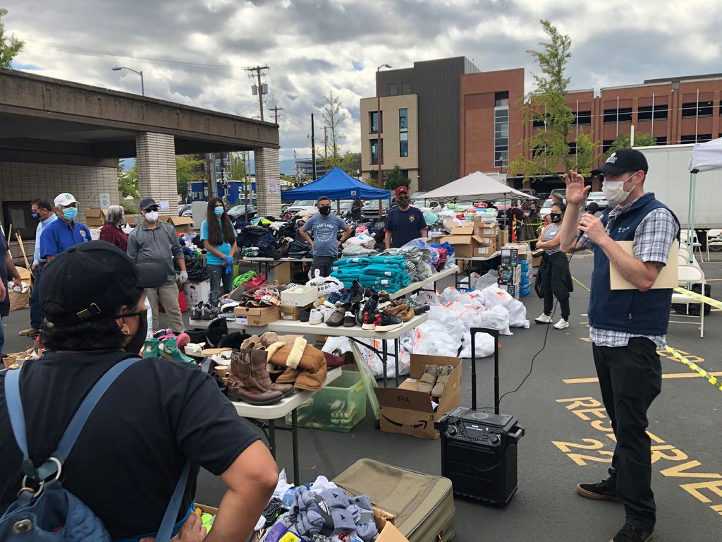 A masked male event coordinator with microphone speaks to crowd of volunteers amid tables of donated clothing.