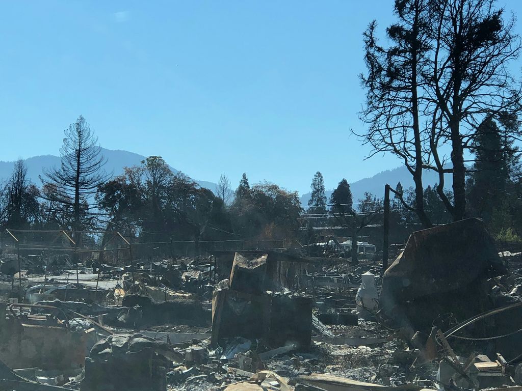 Structures, cars and trees blackened by wildfire, with blue sky and a foothill in the background.