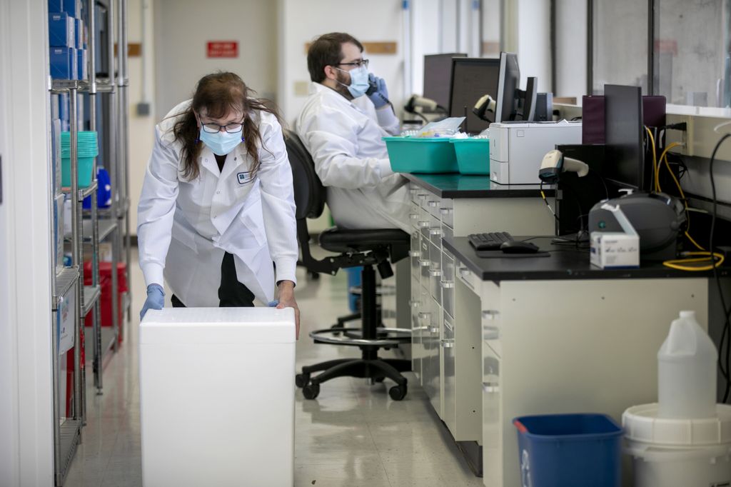 A woman wearing a protective mask pushes a white cube in a research laboratory; a bearded man, also masked, talks on the phone in the background.