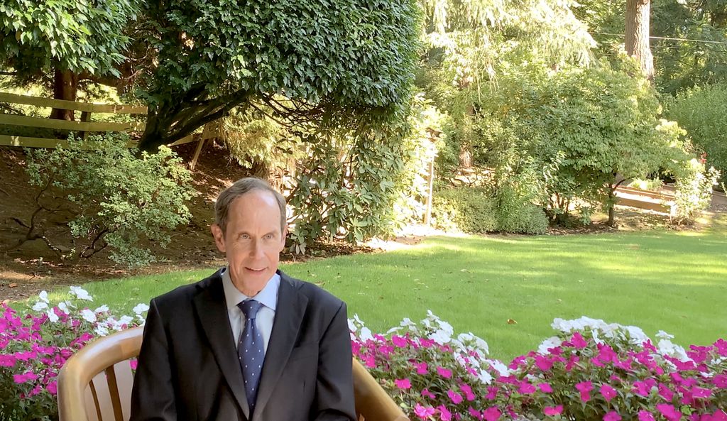 Brian Druker, M.D., a smiling white man in a suit, sits in a wooden chair with a garden in the background.