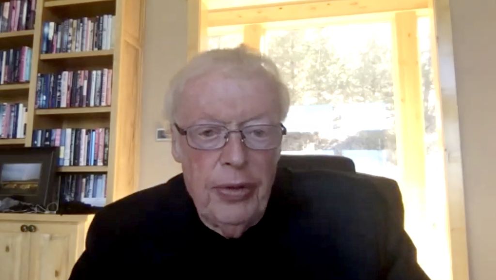 Phil Knight, a caucasian man with white hair and glasses, is shown with bookshelves and a window behind him.