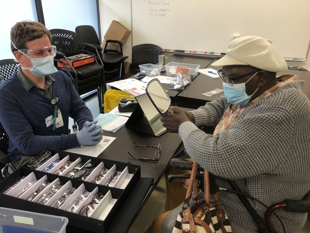 A masked Black woman in a white hat tries on glasses while looking in a hand mirror, across the table from a masked white male volunteer.