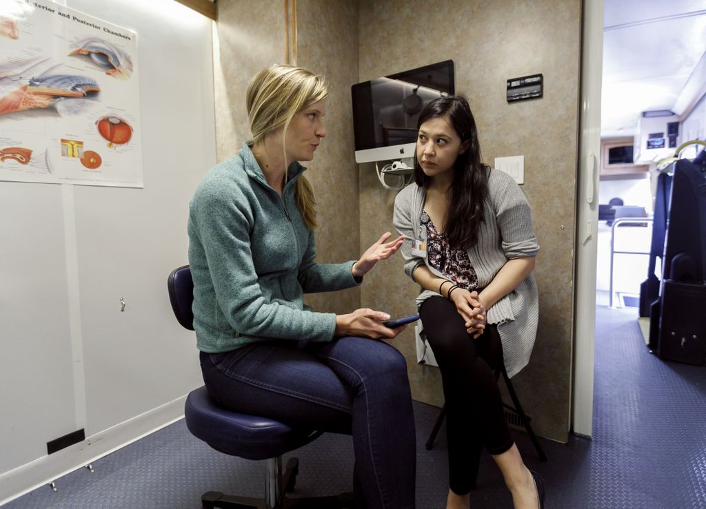 A blond, white woman (left) sits on a stool in a mobile medical office holding a mobile phone, while making eye contact and conversing with a dark-haired female (right), also seated. 