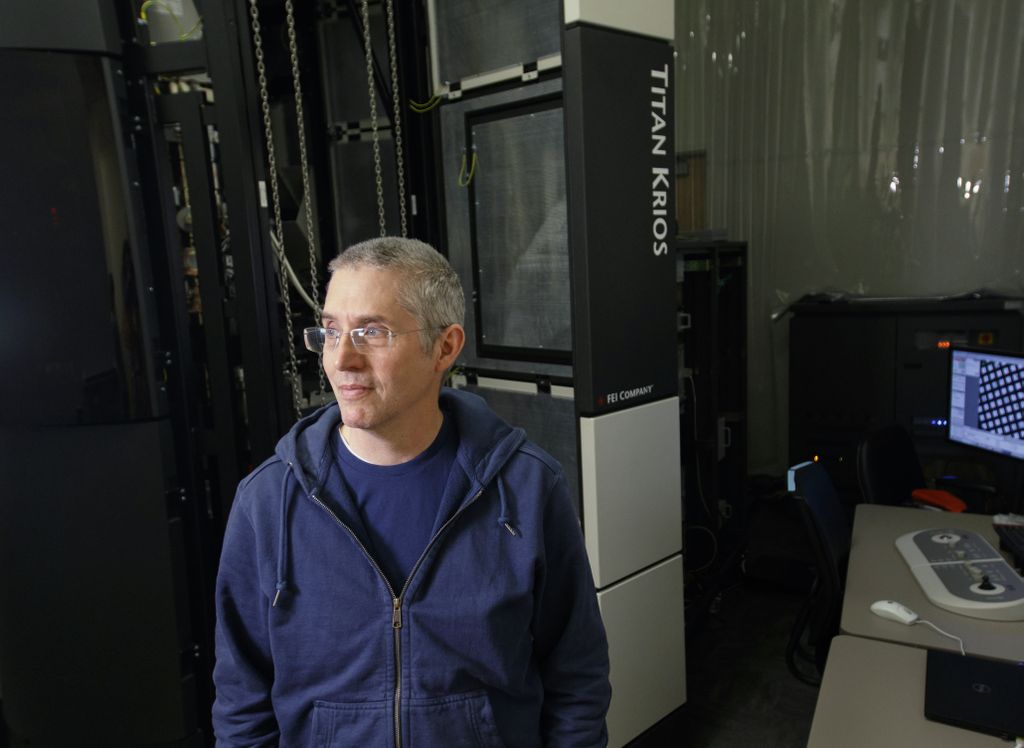 Eric Gouaux, Ph.D., a white man with short grey hair and glasses, stands in front of cryo-EM lab equipment.