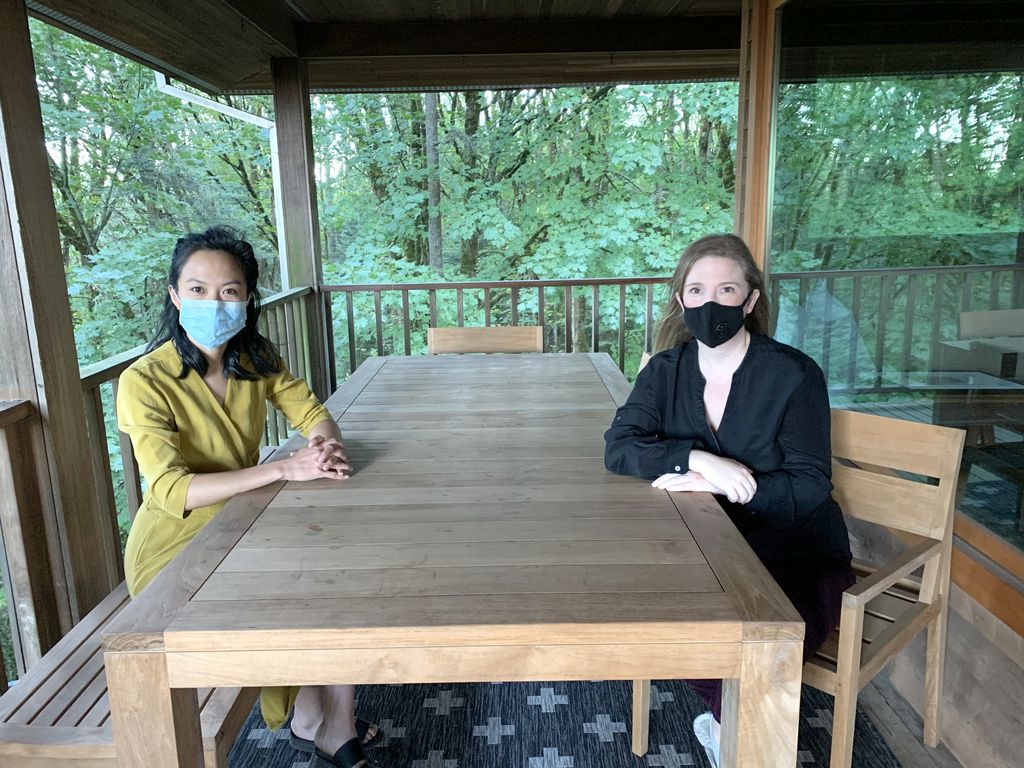 Founders of Urologists for equity, masked females Dr. Geolani Dy (left) and Dr. Casey Seideman (right), sit across from each other at a wooden table with trees visible through the large exterior windows.