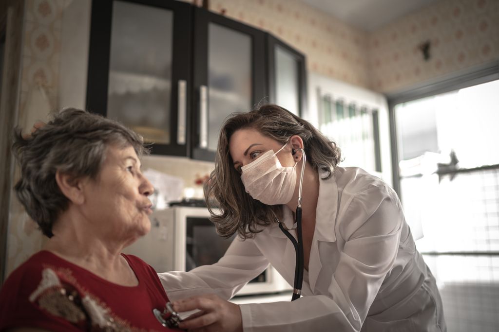 A masked female health care provider in a white lab coat checks a senior woman with a stethoscope during a home visit.