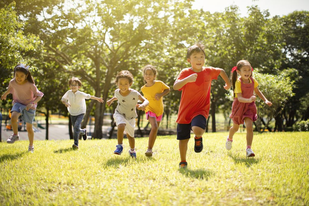 Multi-ethnic group of school children laughing and running on grass with trees and sun in the background.