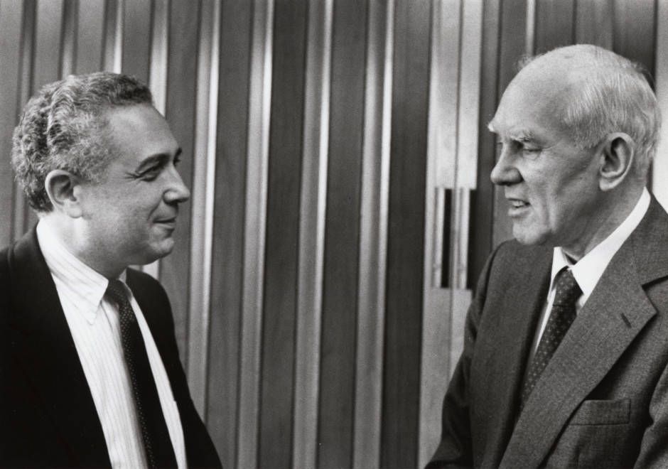 A black and white photo of Leonard Laster and Howard Vollum, two white men in suits, speaking in front of an accordion partition.