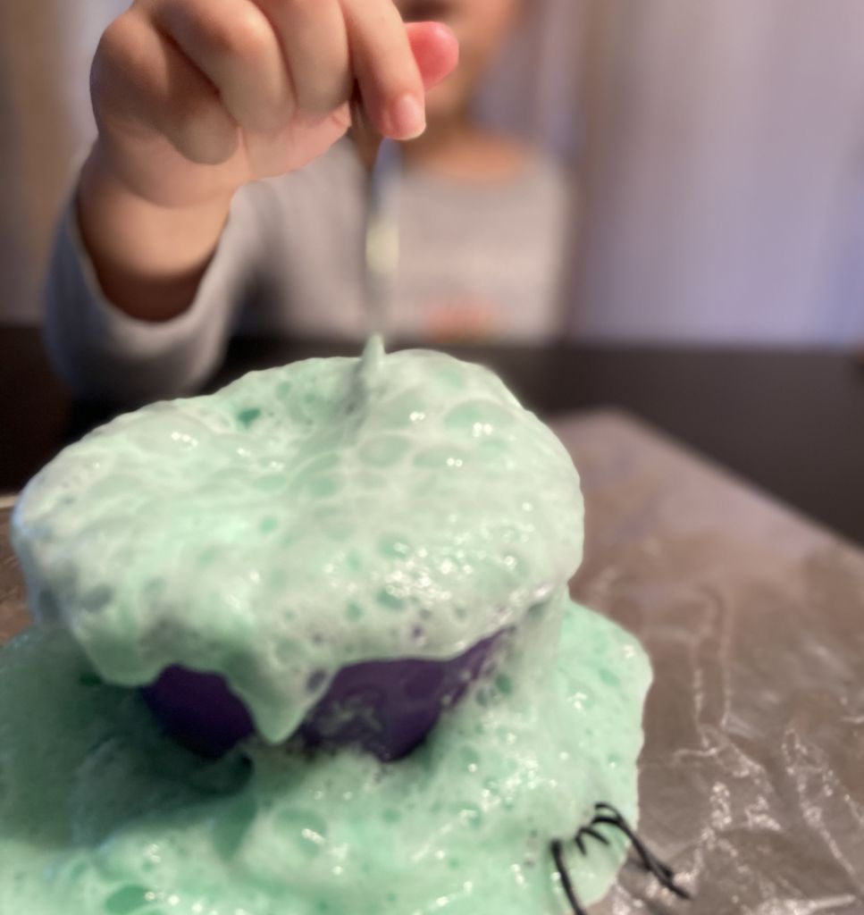 Closeup shot of a small container of "Witches' Brew," a homemade green slime, with a child's hand stirring the mixture.