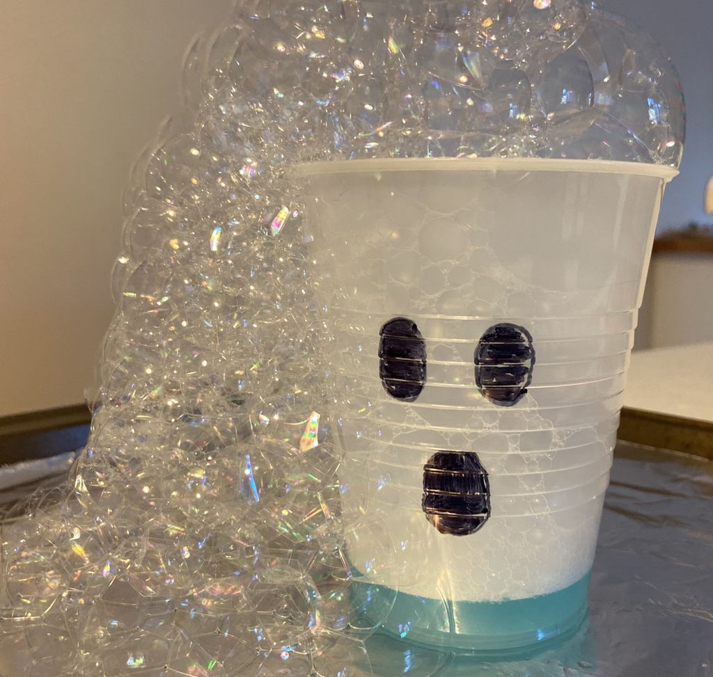 A closeup shot of a white plastic cup with oblong eyes and mouth drawn in black pen, with a foamy bubble solution erupting from the top and spilling over the side.