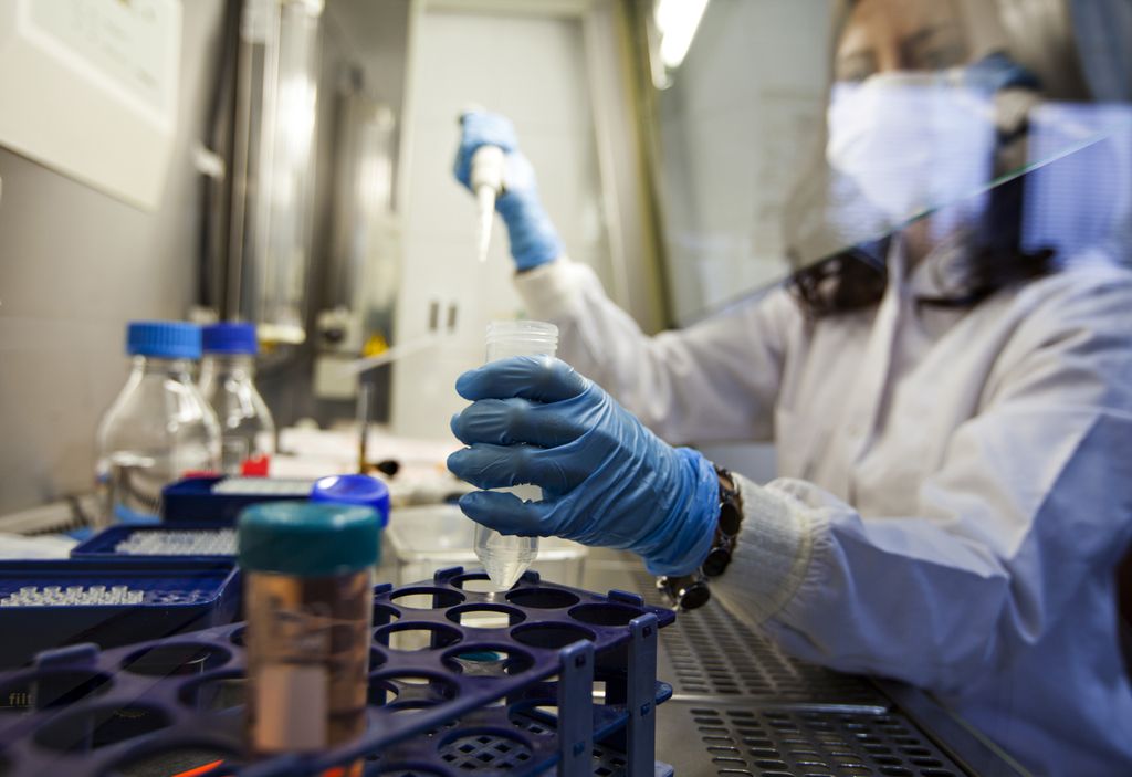 A masked female scientist in blue protective gloves transfers liquid to a vial from a dropper, in a laboratory.