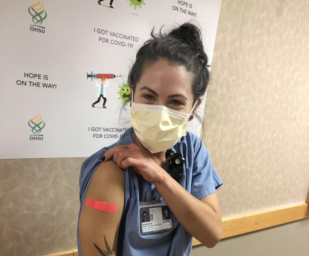 A dark-haired, masked woman pulls up her tshirt sleeve to show the fluorescent pink Band-aid at the site of her COVID-19 vaccine injection on her right shoulder
