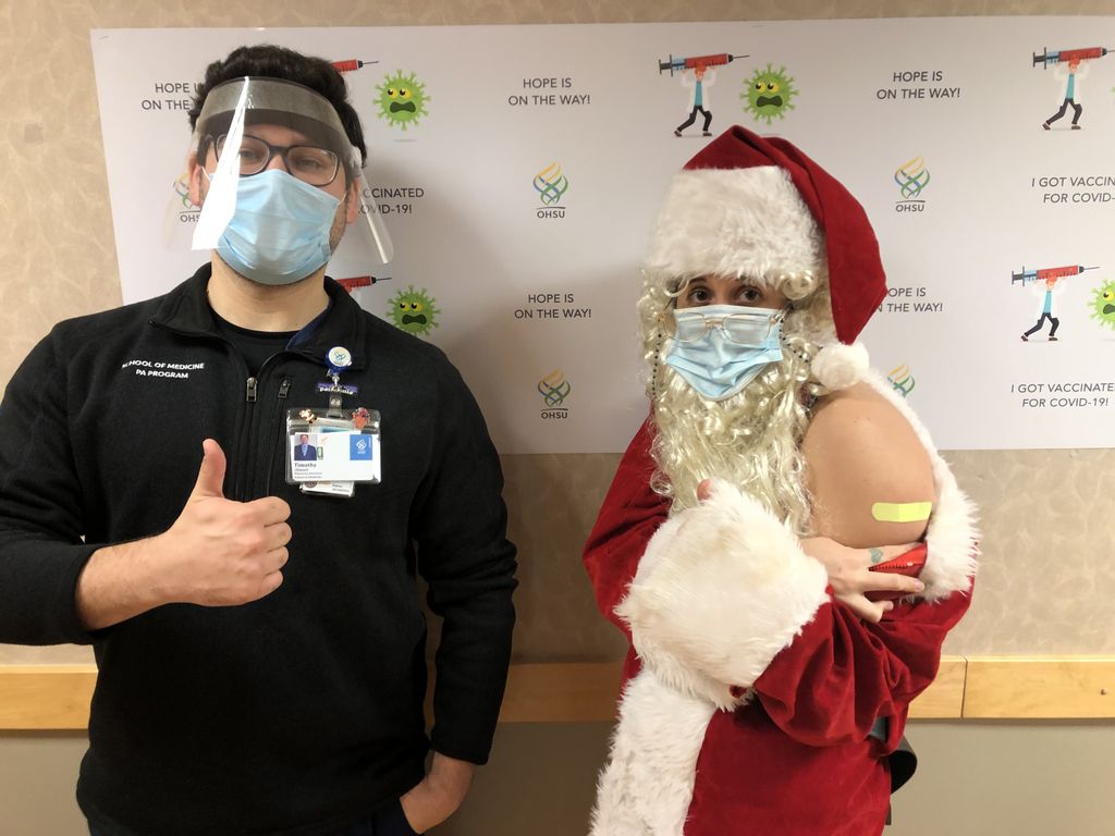 A male Physician's Assistant student (left) wearing a surgical mask and face shield stands giving a thumbs up. A woman in a Santa suit, white costume beard and sugrical mask shows the Band-aid on her shoulder where she received her COVID-19 vaccination shot