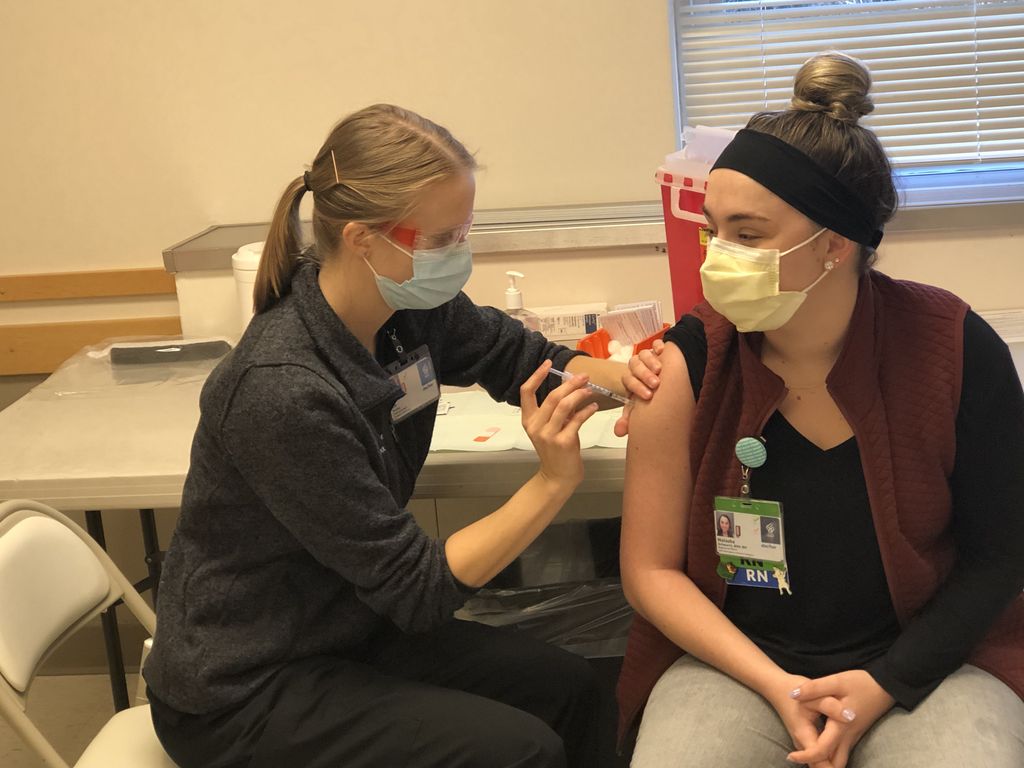 Blonde female in face mask and ponytail injects COVID-19 vaccine into shoulder of another masked caucasian woman.