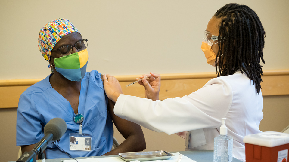 A Black woman in a face mask and scrubs receives a vaccine injection from a dreadlocked dental student in a white lab coat