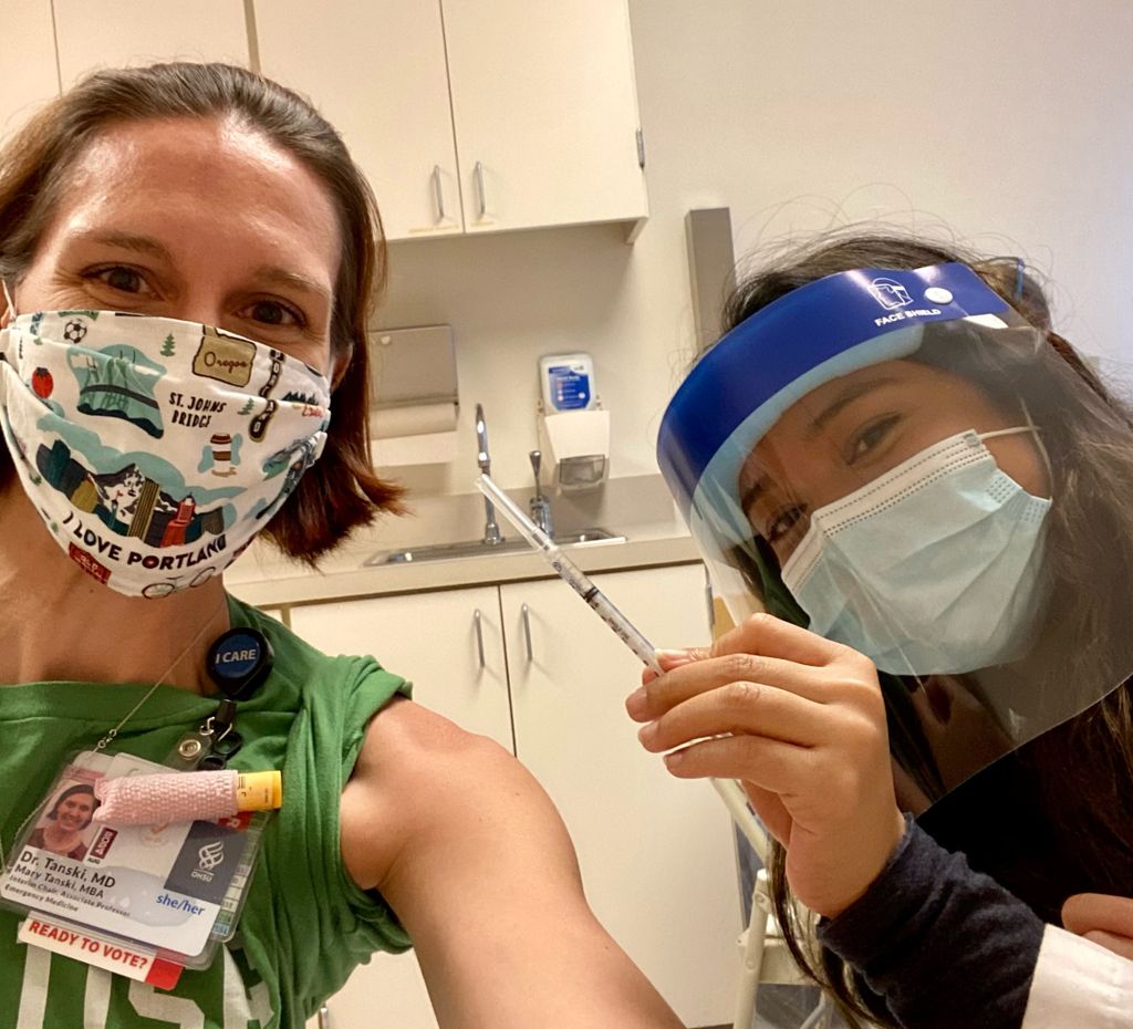 A lean caucasian woman in a face mask offers her shoulder to a masked and shielded Asian female who is holding a syringe of COVID-19 vaccine