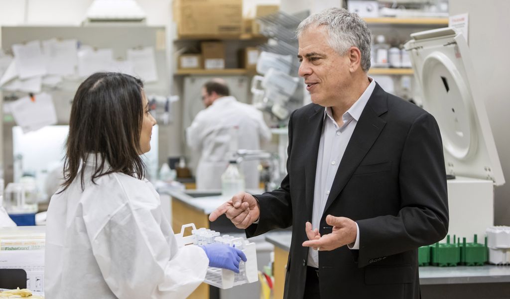 Two adults stand talking in a laboratory; the Asian female (left) is shot in profile, wearing a white lab coat and has dark hair; the white male (right) has grey hair and wears a dark blazer