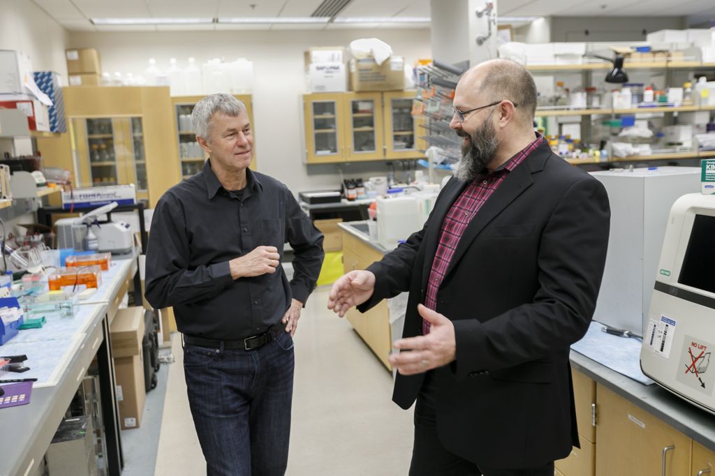 Two caucasian males stand talking in a medical lab; the clean-shaven man on the left has white hair and wears a black button-down shirt; the bearded, bald man on the right wears a plaid shirt and black blazer; medical and lab equipment are around them and in the background