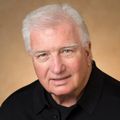 Close-in headshot of an older white male with white hair and a ruddy complexion, wearing a black button-down shirt
