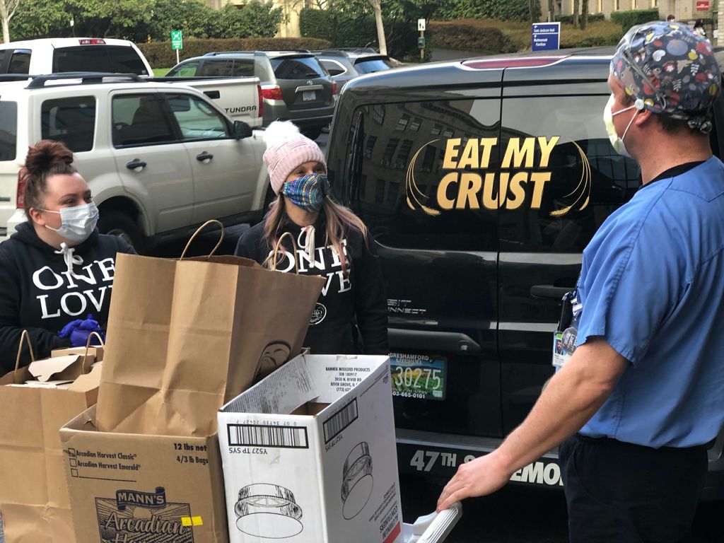 Two masked female adults bring bags of pasta meals to a masked OHSU employee in scrubs and a mask, where he is loading them onto a white wheeled cart