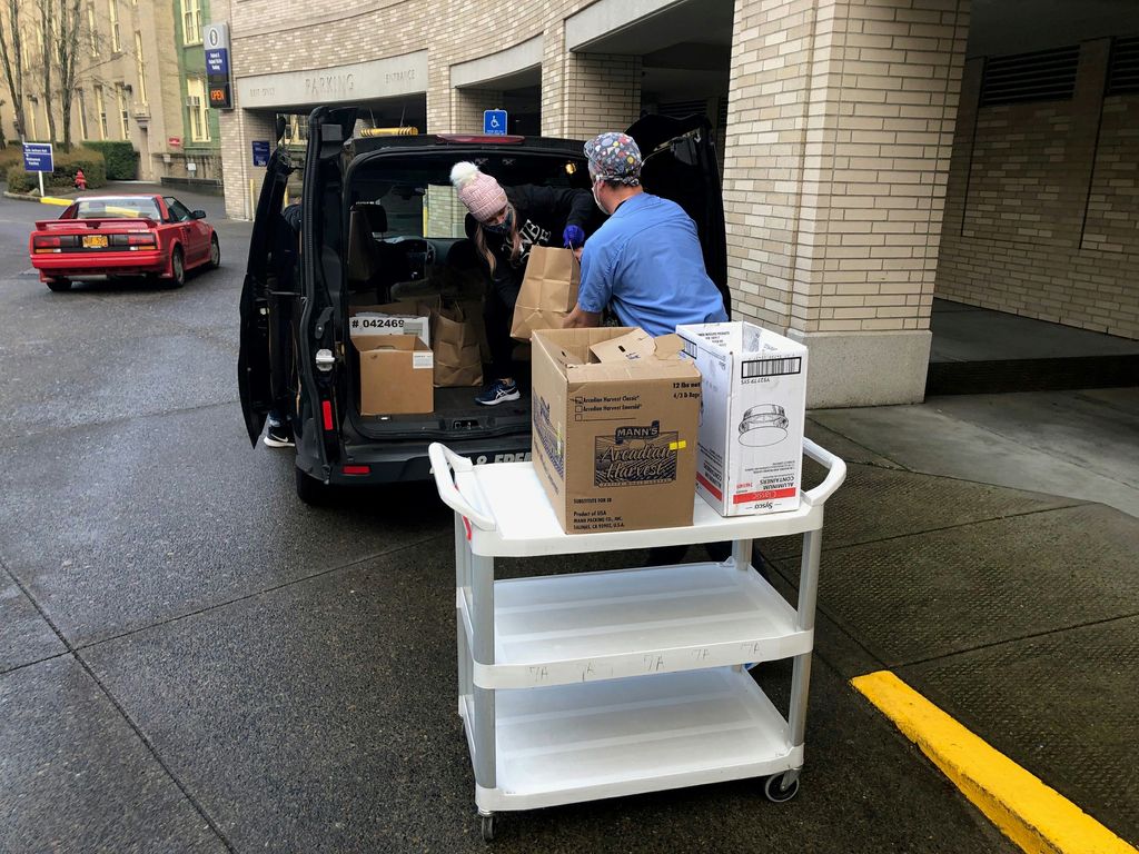 A masked blonde woman in a black beanie hands bags of pasta meals from the back of a delivery van to a masked adult in scrubs, who is placing the bags on a white rolling cart.