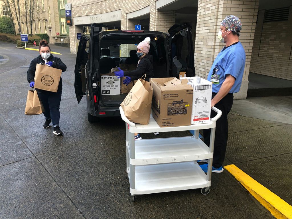 Two masked females bring brown bags of pasta meals to a masked OHSU employee in scrubs, who is loading them onto a white wheeled cart