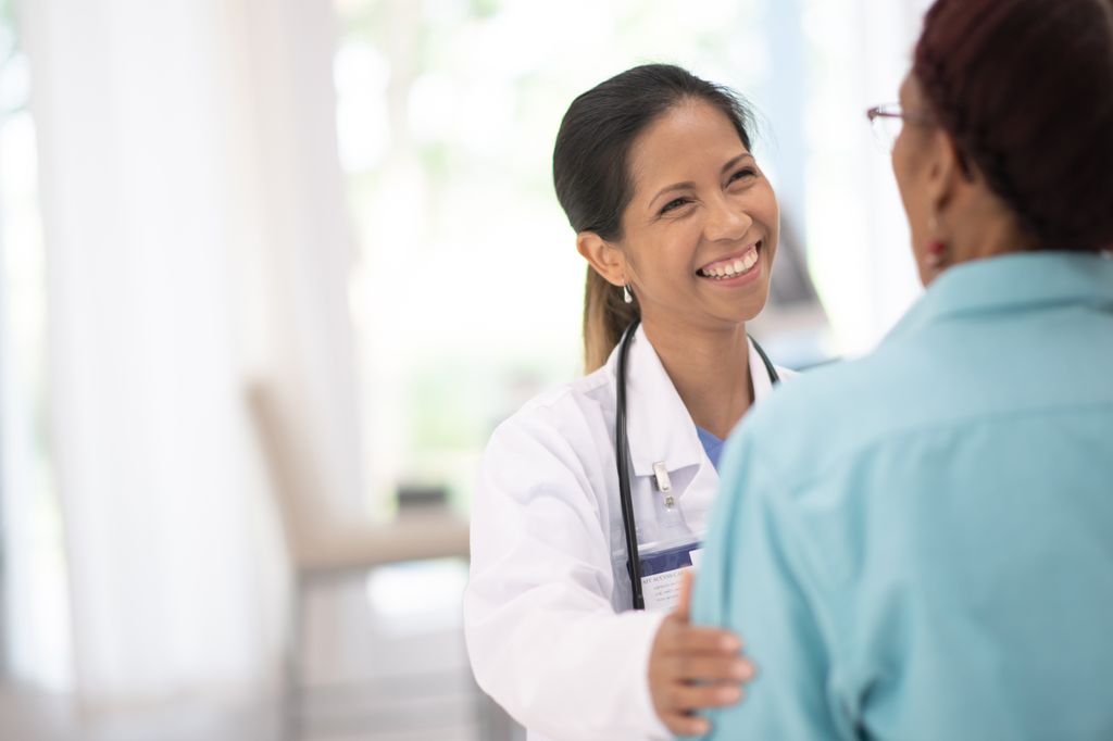 A female doctor wearing a lab coat and stethoscope puts her right hand on the left arm of a female patient, who is shown from behind