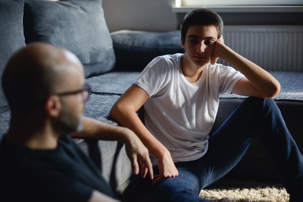 An adult and an adolescent sit on the floor of a living room. They appear to be engaged in a serious conversation. 