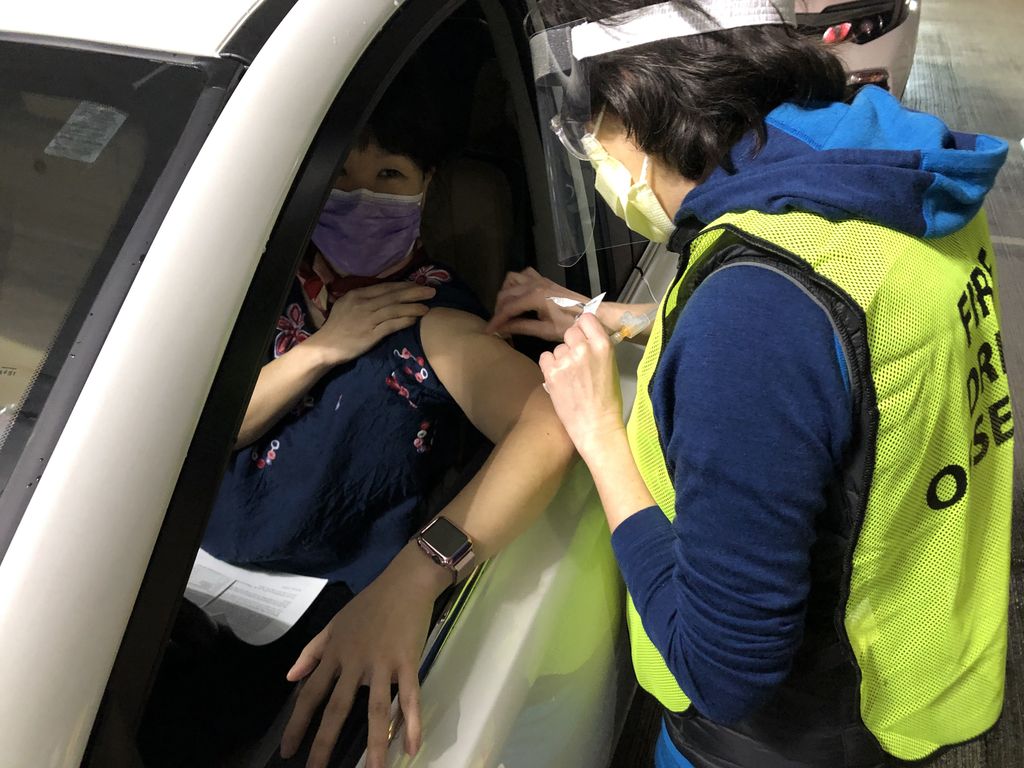 masked health care worker gives injection into the shoulder of a woman in the driver seat of a car