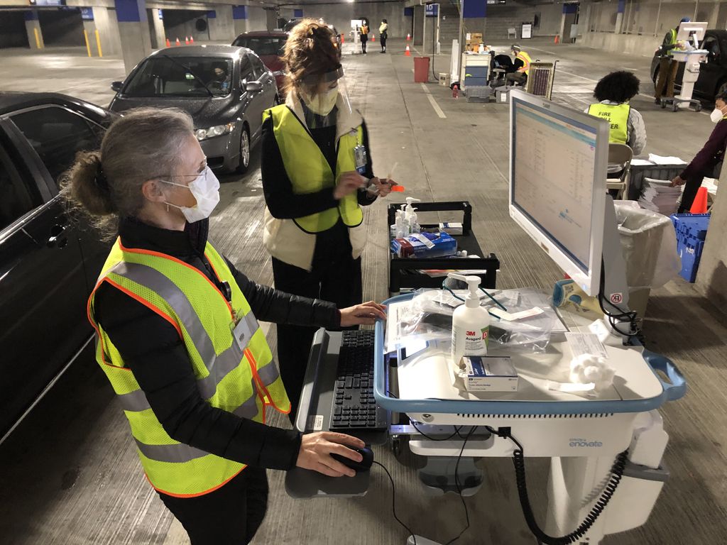 An OHSU doctor and an OHSU nurse in masks and safety vests prepare to vaccinate a caregiver at a drive-up vaccine clinic in the Oregon Convention Center parking garage. Doctor checking the patient's record on a portable computer; nurse is preparing the vaccine.