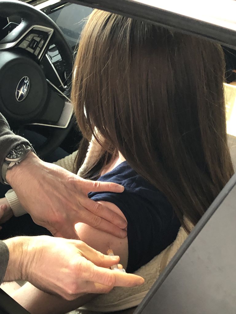 Woman sitting in driver’s side of vehicle receives a shot in her left arm.